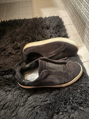 Svarta sneakers från Axel Arigato - Svarta sneakers från Axel Arigato i mocka med elastiska paneler istället för snörning. Snygg minimalistisk design med diskret guldfärgad logga på sidan och vit sula. Perfekta för en clean och stilren look.