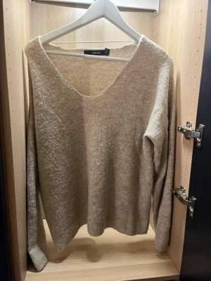 Beige stickad tröja Vero Moda - Mysig beige stickad tröja från Vero Moda med djup v-ringning och långa ärmar. Tröjan har en avslappnad passform och är perfekt för lager-på-lager. Materialet är mjukt och ribbstickat för extra komfort.