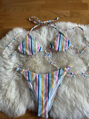 Färgglad randig bikini med knyt - Snygg bikini med färgglada ränder i blått, rosa, gult och vitt. Överdelen har trekantskupor och knyt både i nacken och ryggen. Underdelen har höga ben och knyt i sidorna. Perfekt för stranden eller poolen!