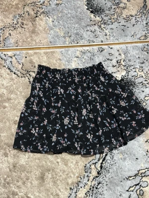 Svart blommig kjol med volanger - Snygg svart kjol med blommigt mönster i multifärg. Har aldrig använts. 