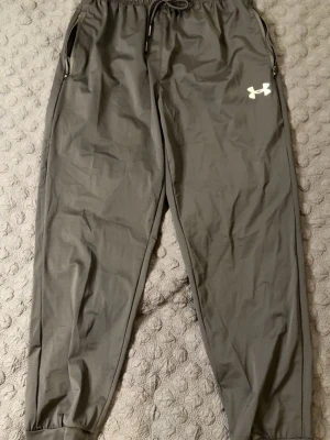 Grå mjukisbyxor från Under Armour - Snygga grå mjukisbyxor från Under Armour med resår i midjan och dragsko. Byxorna har två fickor med dragkedja och en vit logga på vänster lår. Perfekta för chill eller träning, tillverkade i ett lätt och mjukt material. PASSAR M OCH L. HELT NY, OANVÄND