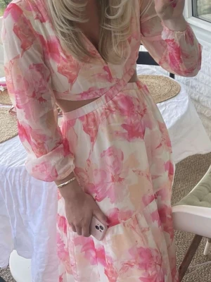 Blommig långklänning med cut-out - Superfin långklänning från Amisu med blommigt mönster i rosa och beige toner. Klänningen har v-ringning, långärmade ärmar och snygg cut-out detalj i midjan. Perfekt för sommarens alla tillfällen!