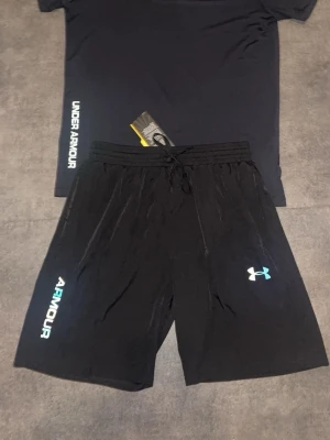 Svarta shorts och t shirt  från Under Armour - Svart sett från Under Armour med logga och text i vitt och blått på vänster ben. Shortsen har snörning i midjan och är tillverkade i ett lätt och bekvämt material, perfekta för träning eller chill. Klassisk passform och sportig look.