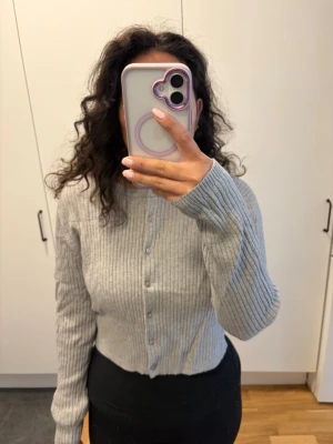 Grå ribbad kofta med knappar - Säljer en grå ribbstickad kofta med knappar framtill. Koftan är croppad och har långa ärmar. Perfekt för lager-på-lager och enkel att matcha med olika outfits.