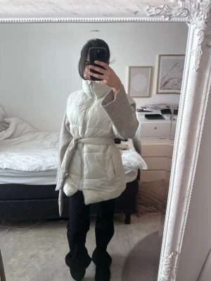 Beige dunväst med knytskärp - Stilren beige dunväst med hög krage och knytskärp i midjan. Västen har två stora fickor framtill och quiltad design som ger en modern look. Perfekt att slänga över en stickad tröja för extra värme och stil.
