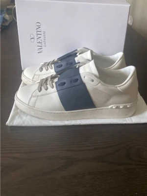 Valentino Garavani vita sneakers - Snygga vita sneakers från Valentino Garavani med en bred mörkblå rem över mitten och matchande blå detaljer vid snörningen. Skorna är i läder och har en klassisk låg siluett med diskreta nitar på hälen. Perfekta för en stilren och trendig look.