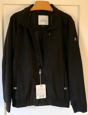 Svart vindjacka från Moncler - Snygg svart vindjacka från Moncler med dragkedja framtill, ribbade muddar och två fickor med dragkedja. Jackan har en stilren design med Moncler-logga på ärmen och är tillverkad i ett lätt, vindtätt material.