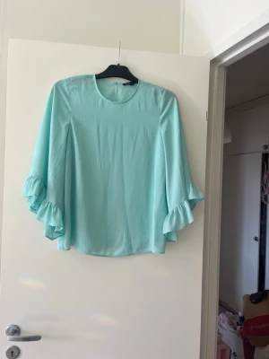 Ljusblå blus med volangärmar - Supersöt ljusblå blus med vida, långa ärmar och volangdetaljer längst ner på ärmarna. Blusen har rund halsringning och är gjord i ett tunt, flowy material som ger en luftig känsla. Perfekt för dig som gillar en feminin och trendig stil.