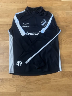 Svart Nike träningströja AIK (half zip) - Svart långärmad träningströja från Nike med AIK-logga på bröstet och vita detaljer längs ärmarna. Halv dragkedja framtill, tryckta sponsorloggor och siffran 49 på framsidan. Perfekt för fotbollsträning eller uppvärmning.