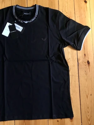 Svart Emporio Armani t-shirt - Snygg svart t-shirt från Emporio Armani med logga broderad på bröstet och märkets namn på kragen. T-shirten har korta ärmar och kontrastdetaljer i vitt på krage och ärmslut. Tillverkad i mjuk bomull för en bekväm känsla.