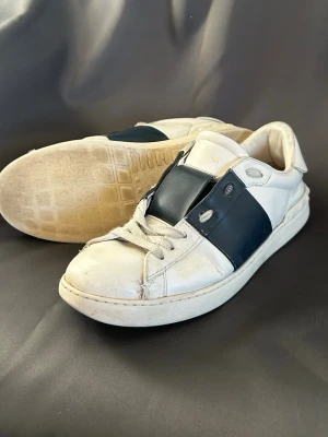 Vit och marinblå Valentino sneaker - Snygga sneakers från Valentino i vitt läder med en bred marinblå läderdetalj över vristen. Klassisk låg modell med rund tå och vita snören. Perfekta för dig som gillar stilrena och exklusiva sneakers med en twist.