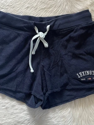 Mörkblå Lexington shorts med snörning i storlek S - Mysiga mörkblå shorts från Lexington i mjuk frotté med bred resårmidja och vit dragsko. Storlek S. Broderad Lexington-logga och flaggor på ena benet. Perfekta för chill dagar eller strandhäng.