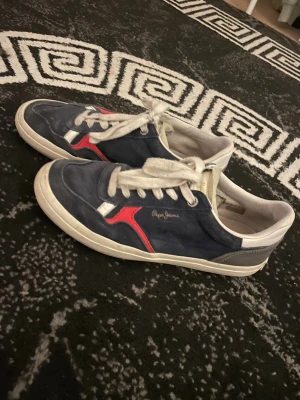 Blåa sneakers från Pepe Jeans - Snygga blå sneakers från Pepe Jeans med vit sula, vita skosnören och detaljer i rött och grått. Ovandelen är i mocka och textil, med logga på sidan. Perfekta för en avslappnad och trendig stil.