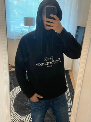 Svart Peak Performance hoodie - Svart hoodie från Peak Performance med broderad vit logga och text 'Est. 1986' på bröstet. Tillverkad i mjukt teddy-material, har huva med dragsko och en stor magficka. Perfekt för en chill och stilren look.
