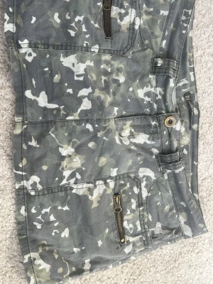 Camouflage kortkjol med dragkedjor - Snygg kortkjol från Trf collection i grönt camouflage-mönster. Kjolen har två framfickor med dragkedjor och två bakfickor. Perfekt för en cool och avslappnad stil. Gjord i bomull för en bekväm känsla. Väldigt fin sommarkväll!