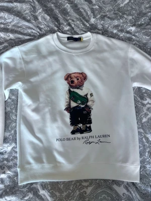 Vit Polo Bear sweatshirt Ralph Lauren - Säljer en vit sweatshirt från Ralph Lauren med den ikoniska Polo Bear-trycket framtill. Tröjan har rund halsringning, långa ärmar och ribbade muddar. Materialet är mjukt och bekvämt, perfekt för en avslappnad stil. Klassisk och stilren design med snygg detalj framtill.