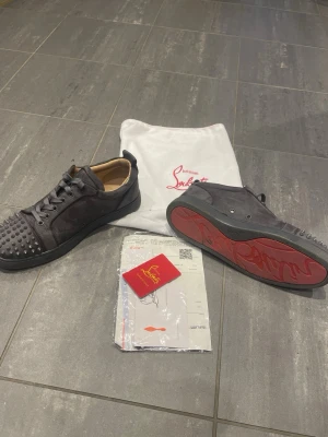 Christian Louboutin gråa sneakers - Snygga Gråa sneakers från Christian Louboutin med klassiska röda sulor och coola nitar på tån. Skorna har lågt skaft, snörning och är tillverkade i mocka. Perfekta för dig som vill sticka ut med en lyxig och edgy stil.