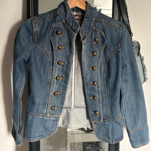 Blå jeansjacka från Denim Co - Snygg blå jeansjacka från Denim Co med militärinspirerad design och dekorativa bronsfärgade knappar framtill. Figurnära passform och coola sömdetaljer ger jackan en unik look. Perfekt för dig som vill sticka ut med din outfit till vår och sommar🌺