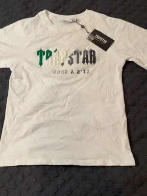 Vit Trapstar t-shirt med broderad logga - Säljer en vit t-shirt från Trapstar med broderad logga i grönt, vitt och grått framtill. T-shirten har rund halsringning och klassisk passform. Perfekt för dig som gillar streetwear och vill sticka ut med snygga detaljer. STORLEK M, PASSAR M-L. HELT NY, OANVÄND.
