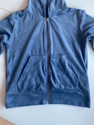 Blå zip-hoodie från 157 - Blå hoodie med dragkedja från lager 157. Modellen har huva, två stora fickor framtill och en snygg ringdetalj på dragkedjan. Perfekt för en avslappnad och stilren look. Mjuk bomullskänsla och klassisk passform i velourtyg. 