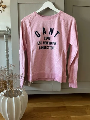 Rosa sweatshirt från GANT - Säljer en ljusrosa sweatshirt från GANT med tryck i mörkblått framtill: 'GANT 1949 EST. NEW HAVEN CONNECTICUT'. Tröjan har rund halsringning, långa ärmar och ribbade muddar. Perfekt för en avslappnad och sportig stil.