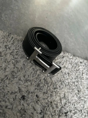 Svart skärp med silverfärgat H-spänne - Snyggt svart bälte med ett stort, silverfärgat H-format spänne. Bältet har vita sömmar längs kanterna som ger en clean kontrast. Perfekt accessoar för att lyfta vilken outfit som helst.