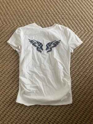Vit t-shirt med vingar från Bershka - Snygg vit t-shirt från Bershka med marinblått tryck av vingar på framsidan. Klassisk passform och rund halsringning. Tillverkad i mjuk bomull som känns skön mot huden. 