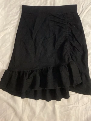 Svart volangkjol med rynkade detaljer - Svart kjol med volangkant och rynkade detaljer på ena sidan. Kjolen har en lätt utsvängd form och är tillverkad i ett mjukt tyg som ger en snygg siluett. Perfekt för dig som gillar en stilren men ändå lekfull look.