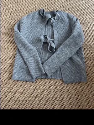 Grå ribbad kofta med knytband - Säljer en stilren grå ribbad kofta med långa ärmar och knytband framtill. Koftan har en rak passform och är tillverkad i ett mjukt stickat material som känns skönt mot huden. Perfekt för lager på lager och enkel att matcha med olika outfits. Man kan knyta om och ta bort rosetterna om man vill.