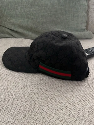 Gucci keps - Snygg svart keps från Gucci med diskret GG-monogram över hela kepsen. På sidan finns en klassisk grön och röd rand. Kepsen har böjd skärm och justerbar rem bak. Perfekt för dig som gillar exklusiva accessoarer.