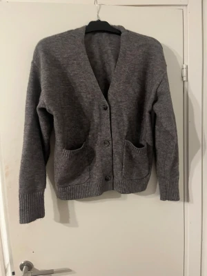 Grå stickad kofta med fickor - Mysig grå stickad kofta med v-ringning och knäppning framtill. Två praktiska fickor på framsidan och ribbade muddar vid ärmslut och nederkant. Perfekt för lager på lager och enkel att matcha med olika outfits.