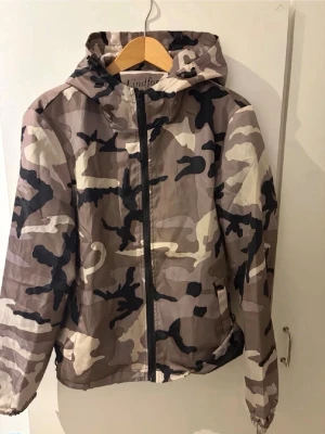 Camo vindjacka med huva - Snygg vindjacka från Lindfors i beige, brunt och svart camomönster. Jackan har dragkedja framtill, två sidofickor och en praktisk huva. Tillverkad i lätt och vindtätt material, perfekt för blåsiga dagar. Vid mer info skriv gärna! ABS! Pris går att diskutera! 