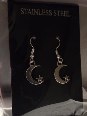 Måne och stjärna örhängen i stål - Snygga örhängen i rostfritt stål med hängande måne och liten stjärna. De är silverfärgade och har en cool, minimalistisk design som passar perfekt till många olika stilar. Krokarna gör dem enkla att ta på och av.