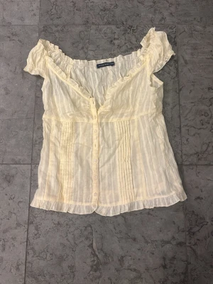 Gulrandig blus med volangkant - Söt gulrandig blus från brandy Melville  med knappar framtill, volangkant vid halsringning och nederkant samt korta ärmar. Tunn och luftig i ett lätt, transparent tyg med diskreta vertikala ränder och dekorativa veck framtill.