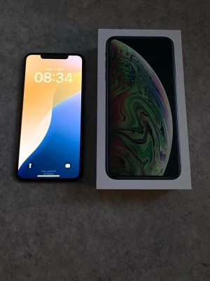 iPhone XS Max 64GB - Säljer en svart iPhone XS Max från Apple med stilren design och glasbaksida. Telefonen har en stor skärm med tunna kanter och Face ID. Baksidan är blank och har Apples logga. Kommer med originalkartong..