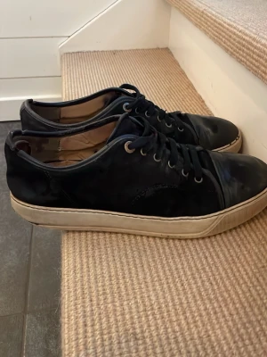 Svarta sneakers från Lanvin - Snygga svarta sneakers från Lanvin med klassisk låg profil och vita sulor. Skorna har ovandel i mocka och läder, svarta skosnören och diskreta sömdetaljer. Perfekta för dig som gillar stilrena och tidlösa sneakers. Finns lite bekymmer med skorna därav kan pris diskuteras. Ställ frågor om något behöver vetas.