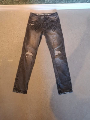 Grå slitna jeans med tryck från hoodrich JDsports - Säljer ett par grå jeans med slitningar och hål på båda benen men det går inte igenom till benen. Perfekta för en streetwear-look.