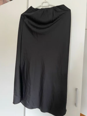 Svart långkjol i satin-look - Stilren svart långkjol med elastisk midja och mjukt, glansigt material som ger en snygg satin-look. 