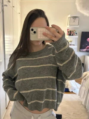 Grå stickad tröja med vita ränder - Mysig grå stickad tröja med horisontella vita ränder. Tröjan har en avslappnad passform, rund halsringning och långa ärmar. Perfekt för lager på lager och enkel att matcha med jeans eller mjukisbyxor.