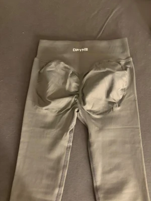 DFYNE Seamless Scrunch Leggings - Khaki / Ljusgrå - Storlek XS - Säljer ett par superfina DFYNE seamless leggings i en unik färg som drar mot både khaki och ljusgrått. Storlek Extra Small (XS). DFYNEs klassiska komfort och kvalitet! De har den populära scrunch butt-effekten som ger en jättefin form. Materialet är squat-proof, stretchigt och har en bekväm hög midja som sitter stadigt på plats. Tightsen är i mycket fint skick och säljes då jag rensar ut i garderoben. Ett perfekt basplagg för gymmet! • Märke: DFYNE • Färg: Khaki / Ljusgrå • Storlek: XS • Skick: 