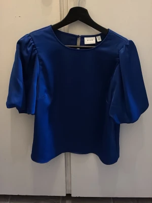 Blå blus från VILA - Snygg blå blus från VILA med puffiga korta ärmar och rund halsringning. Enkel knäppning bak i nacken och rak passform. Perfekt för att lyfta vilken outfit som helst.