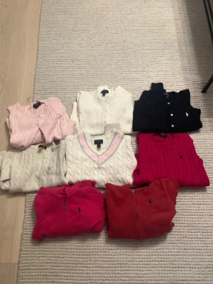 Stickade tröjor från Ralph Lauren - Säljer flera stickade tröjor från Ralph Lauren i olika färger som vitt, rosa, rött, marinblått och cerise. Tröjorna har kabelstickat mönster, vissa med dragkedja och andra med v-ringning eller rund hals. Klassisk Polo-logga broderad på bröstet. Storlek 8-10år