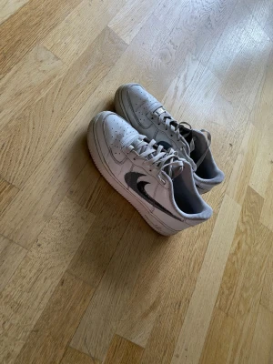 Vita Nike Air Force 1 sneakers - Klassiska vita Nike Air Force 1 sneakers med svart swoosh på sidan. Skorna har lågt skaft, perforerad tåbox och vita snören. Tillverkade i läder med gummisula. Perfekta för en clean och stilren look.