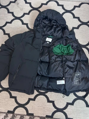 Svart pufferjacka från Lacoste - Säljer en svart pufferjacka från Lacoste med huva och dragkedja. Jackan har grön innerficka, är vattenavvisande och fodrad med återvunnen polyester och dun för extra värme. Perfekt för kalla vinterdagar och har en sportig look med diskret logga på insidan.