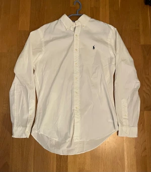 Vit skjorta från Ralph Lauren - Klassisk vit skjorta från Ralph Lauren med button-down krage och broderad logga på bröstet. Perfekt till sommarn Skjortan har lång ärm, knappar framtill och är tillverkad i bomull. Perfekt till en clean och stilren look.