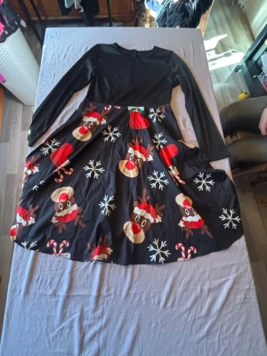 Svart julklänning med renar och snöflingor - Rolig svart klänning med lång ärm och färgglatt julmönster med renar, snöflingor, tomteluvor och polkagrisar. Överdelen är enfärgad svart och kjolen har ett utstickande mönster. Perfekt för julens alla tillfällen.