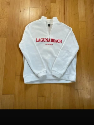 Vit quarter zip sweatshirt Laguna Beach - Vit sweatshirt med quarter zip och trycket 'LAGUNA BEACH CALIFORNIA' i rött på bröstet. Tröjan har hög krage, långa ärmar och ribbade muddar. Materialet är mjukt och bekvämt, perfekt för en avslappnad stil.