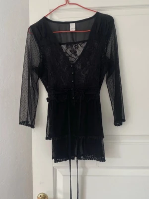 Svart spetskofta med knytband - Snyggt svart kofta i spets med v-ringning och knappar framtill. Trekvartsärmar och dekorativt knytband i midjan. Fina spetsdetaljer och volangkant nedtill ger en romantisk känsla. Perfekt för dig som gillar en elegant och feminin stil.