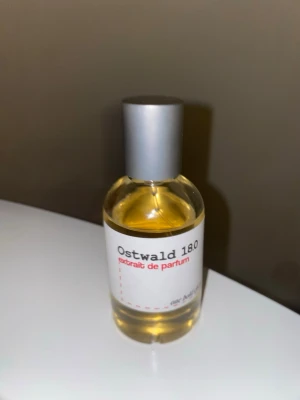 Ostwald 18.0 Extrait de Parfum - Ostwald 18.0 Extrait de Parfum i en rund glasflaska med silverfärgad spraykork. Etiketten är vit med svart och röd text. Parfymen har en ljusgul vätska och minimalistisk design som känns modern och stilren.