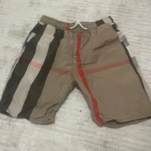 Burberry shorts  - Säljer ett par burberry shorts med beige, svart, vit och röd färg. Shortsen har elastisk midja med snörning och ett klassiskt rutigt mönster. Perfekta för sommaren och har en loose passform.